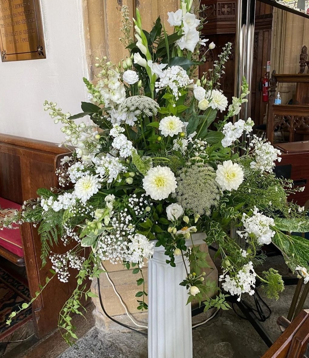 Tall white and green pedestal arrangement of dahlias, stocks, Queen Anne's lace and gypsophila on a white column inside a church