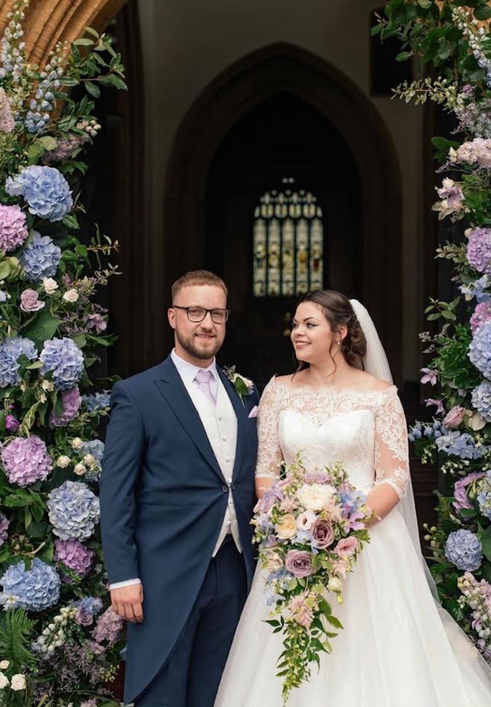 Bride with a trailing pastel bouquet and groom between two tall columns of blue and lilac hydrangeas in a church doorway