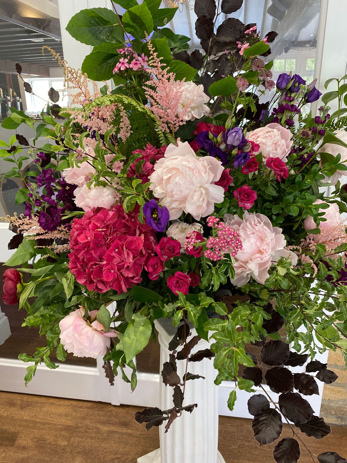Pedestal arrangement of pink peonies, raspberry roses and burgundy foliage