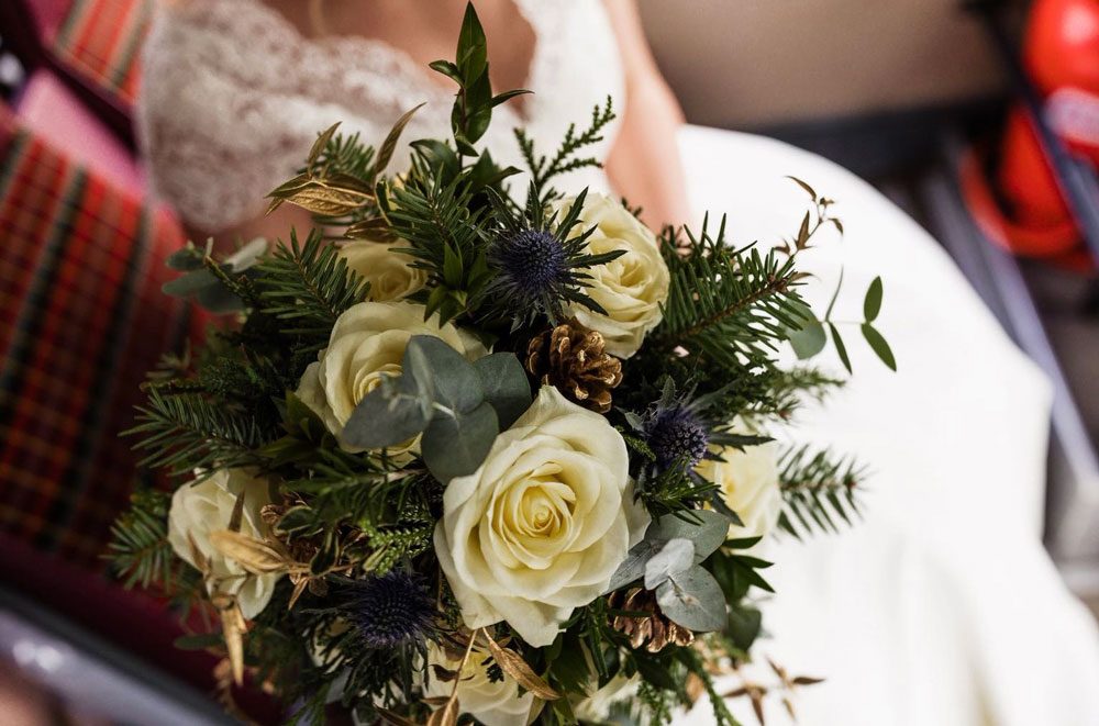 Winter bouquet of ivory roses, blue thistles, pine and pine cones held against a wedding dress